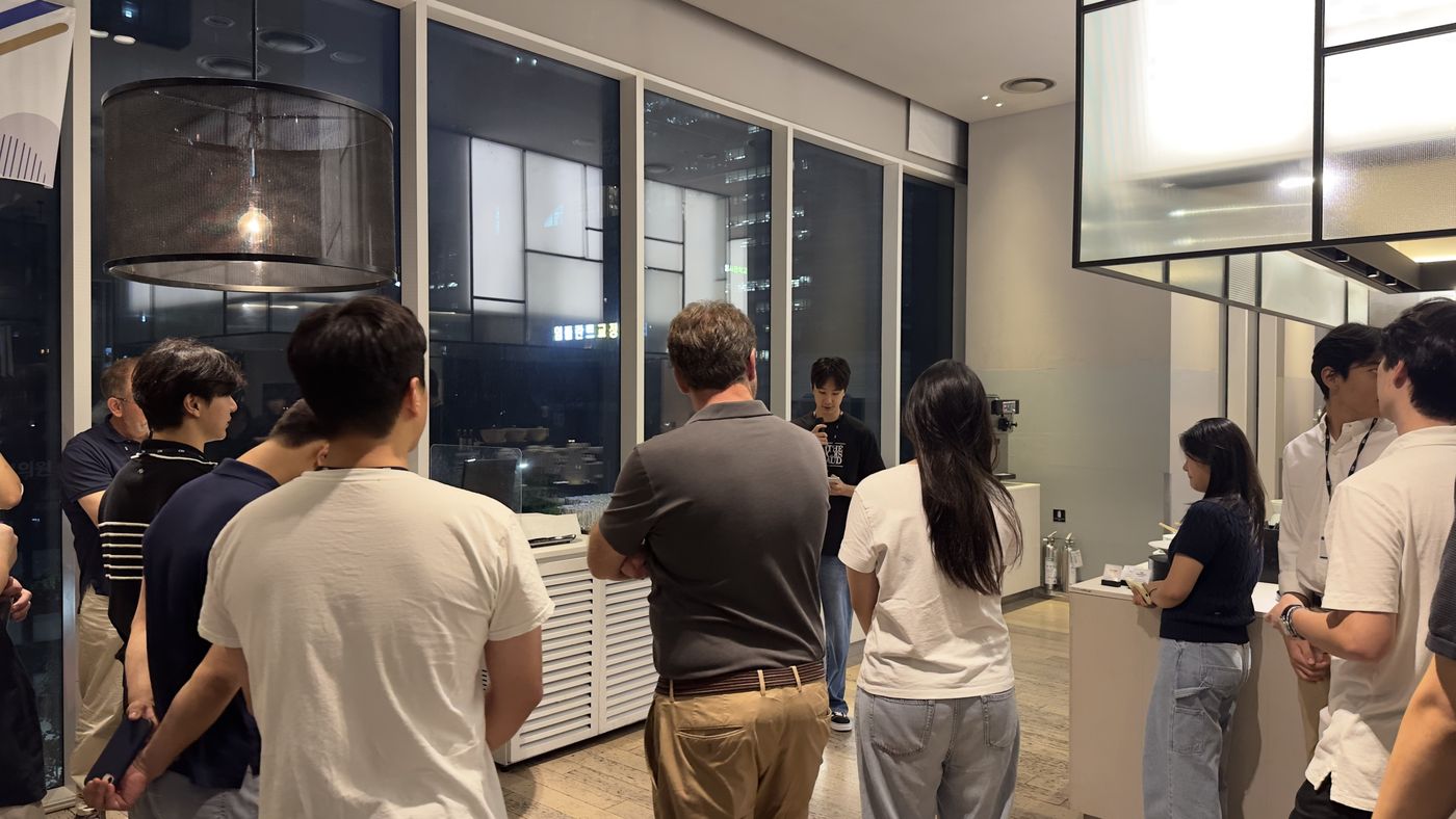 A student holding a microphone speaks at the far end of the room while a group of students and adults stand around listening