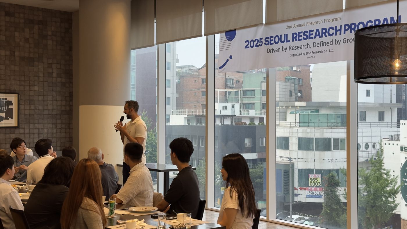A professor in a white polo shirt speaks into a microphone beside tables of seated students