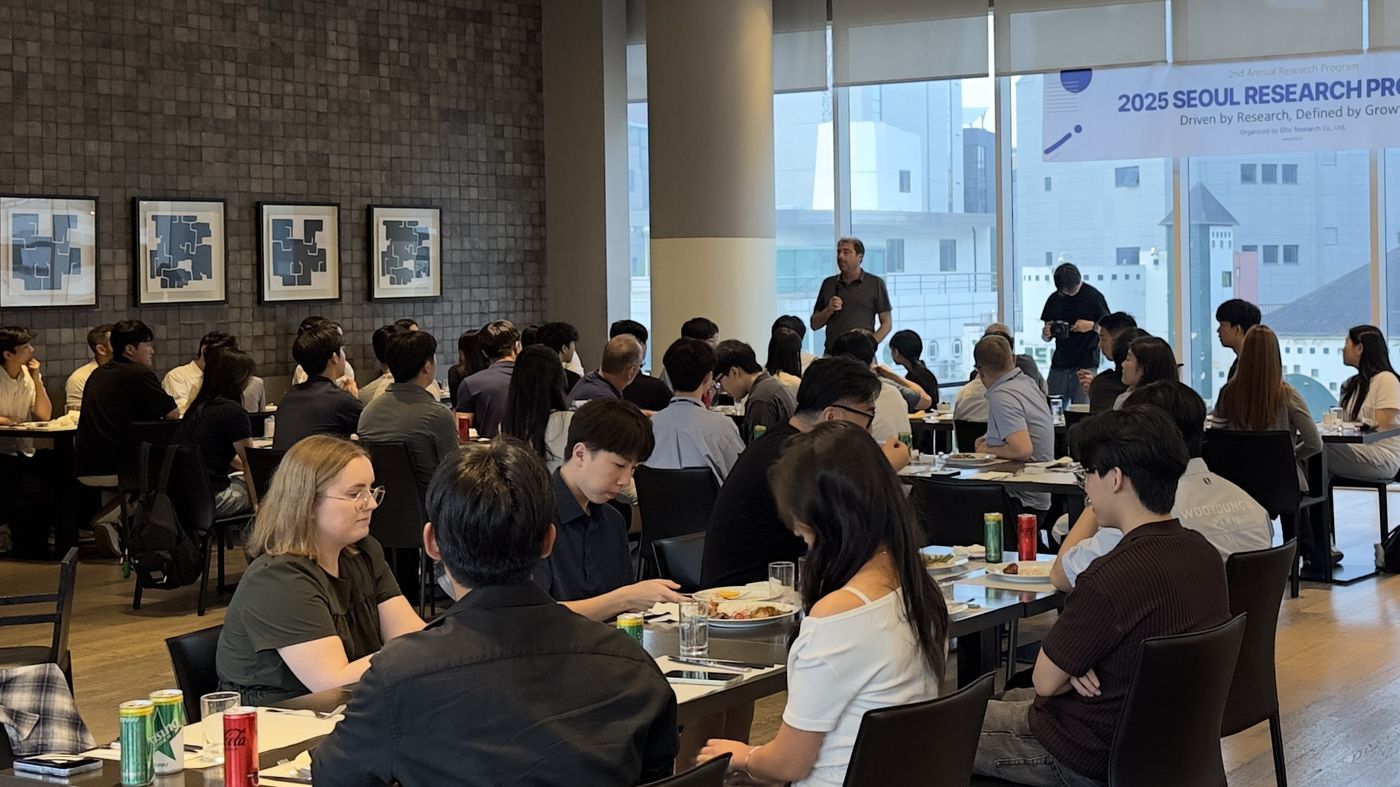 A man stands in the middle of a crowded dining room, speaking to students and faculty seated at long tables, with a 2025 Seoul Research Program banner across the windows