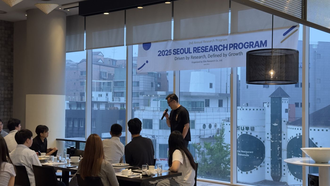 A professor in a black shirt speaks into a microphone in front of floor-to-ceiling windows as students listen from their tables