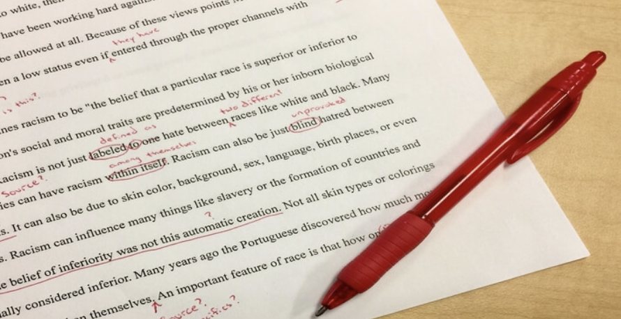 A printed essay covered in red-pen edits and margin notes, with a red pen lying across the page
