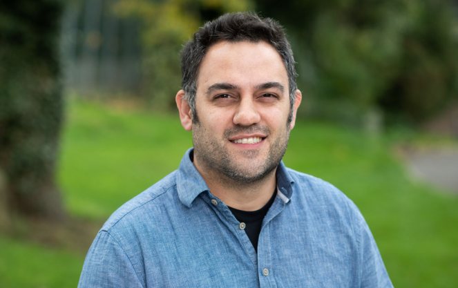 Aziz Aboobaker, smiling, in a blue button-down shirt, standing outdoors in front of a lawn and trees