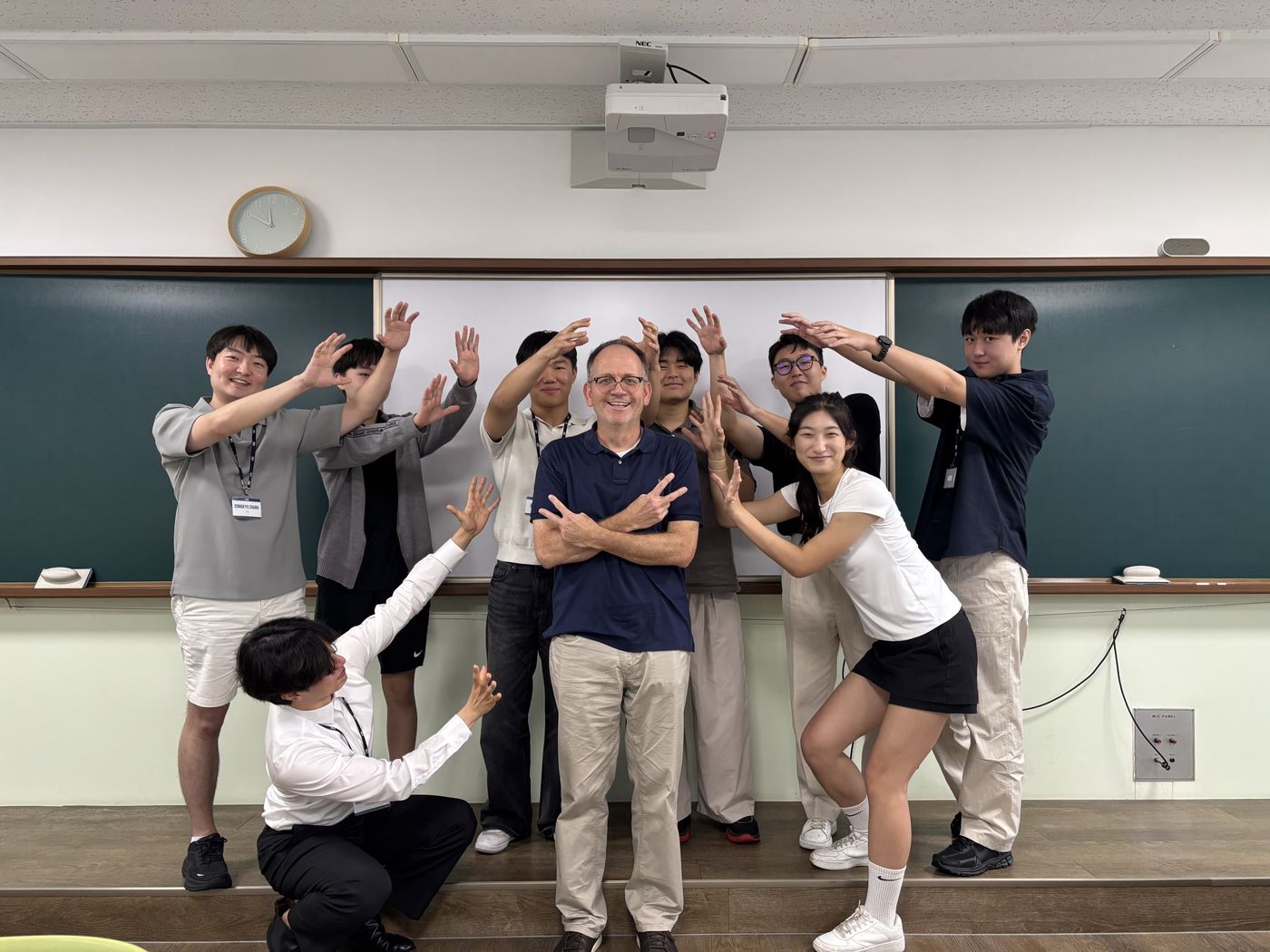 Brian Stoltz stands with arms crossed among about ten students striking playful poses in front of a classroom whiteboard