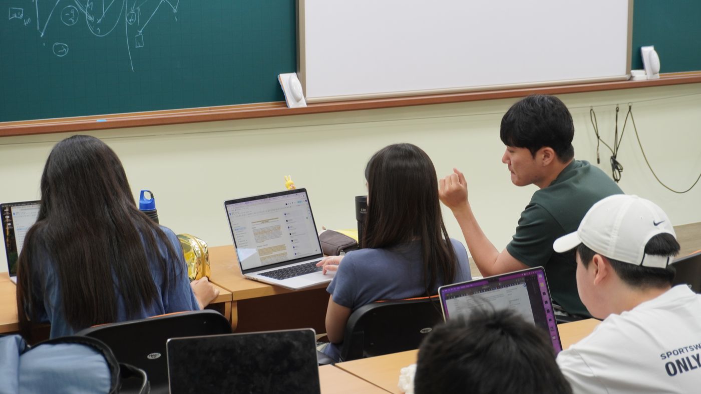 Students work on laptops at classroom desks while an instructor in a green shirt leans in to talk with one of them