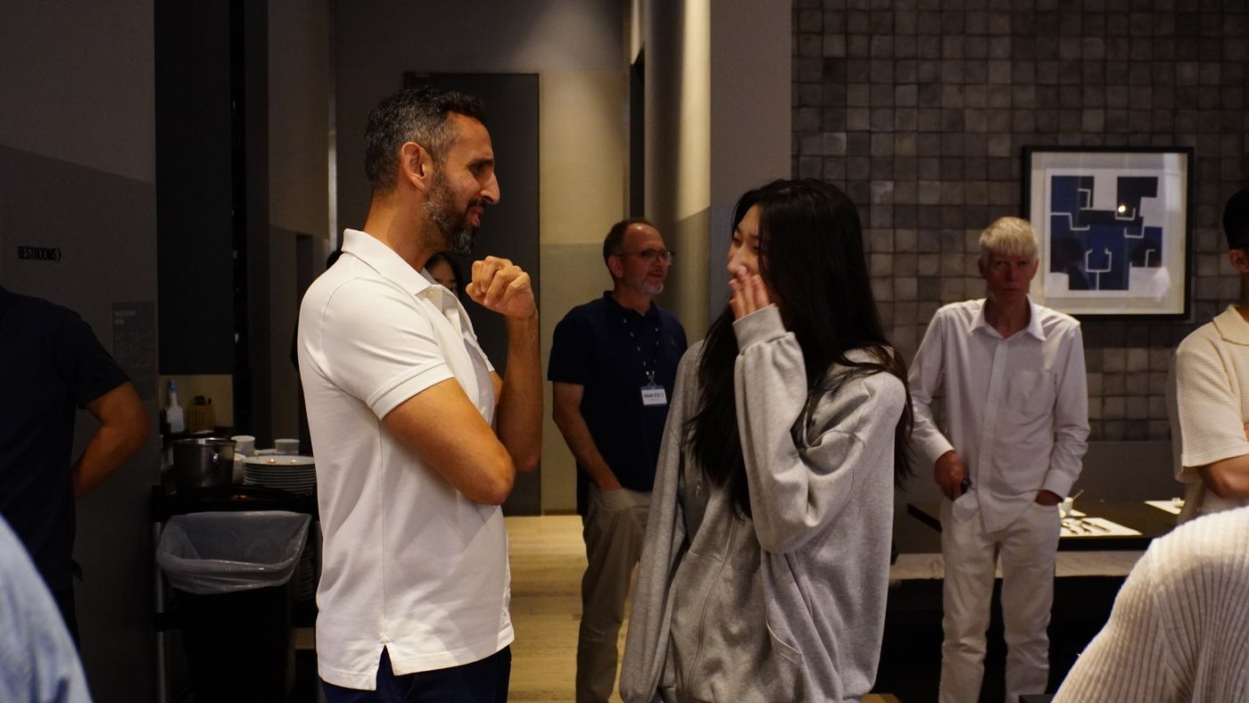 A professor in a white polo shirt talks with a student in a gray hoodie at an evening gathering, with other faculty standing nearby