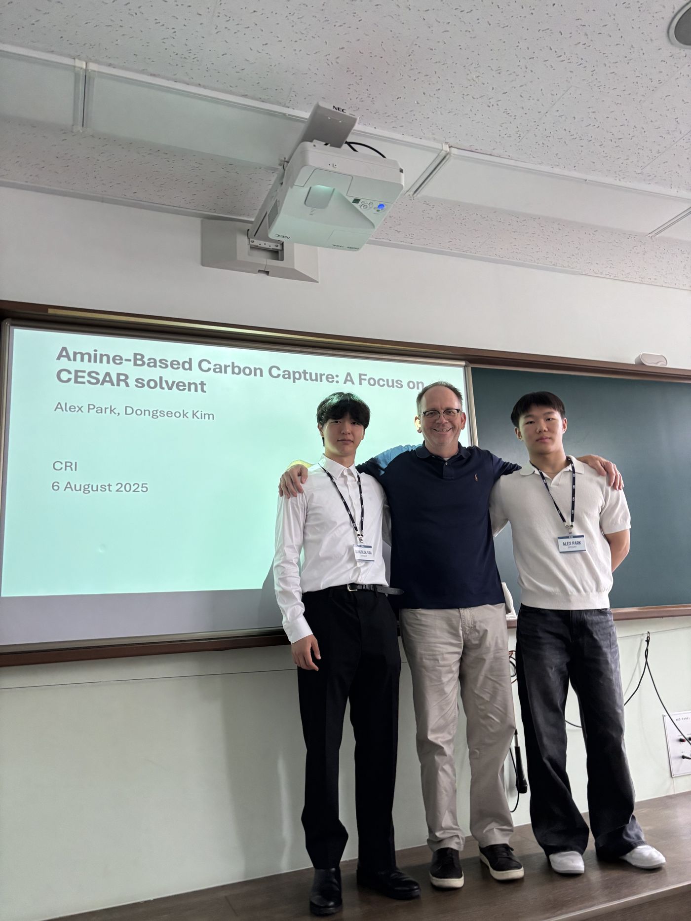 Two students wearing lanyards stand with their professor in front of a projected slide titled "Amine-Based Carbon Capture: A Focus on CESAR solvent"