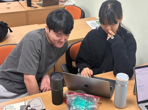 A young man leans in beside a student to look at her laptop screen, with bagged electronic parts on the desk