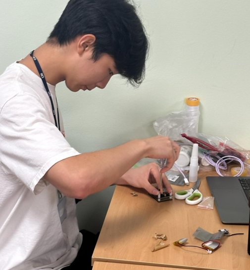 A student leans over a desk, fitting components onto a small circuit board, with wires and a laptop nearby
