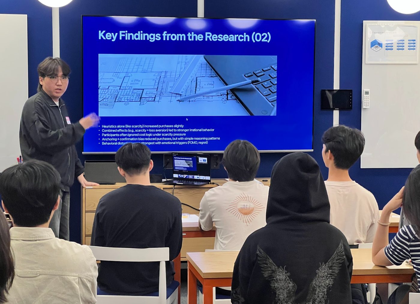 P. stands beside a large screen showing a slide titled "Key Findings from the Research (02)" while students seated at tables listen