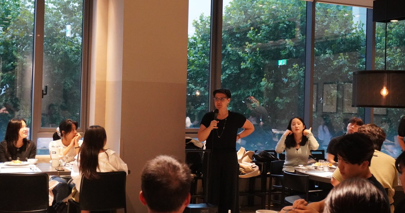 A professor holding a microphone speaks to students seated at dining tables in a room with tall windows