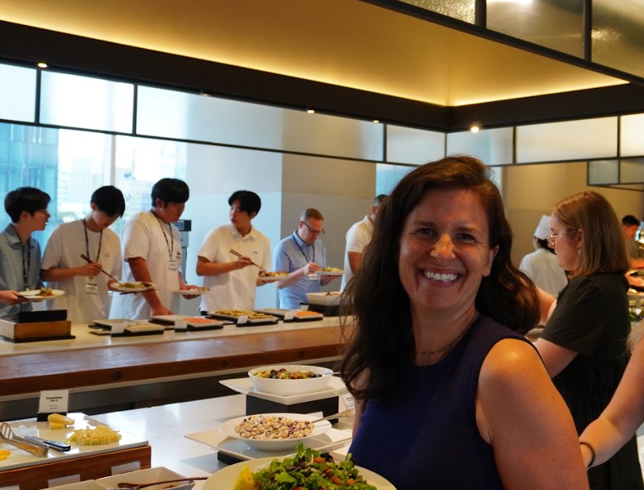 Kit Hickey smiles beside a buffet counter while students wearing lanyards serve themselves in the background