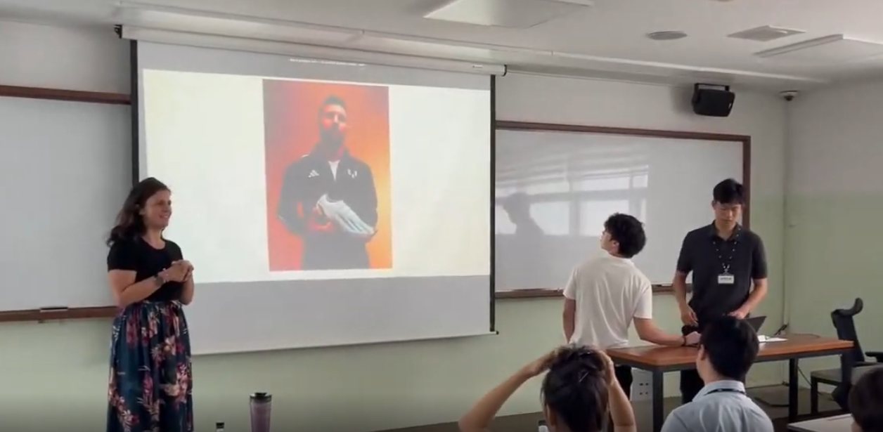 Kit Hickey stands at the front of a classroom as two students present in front of a projected slide