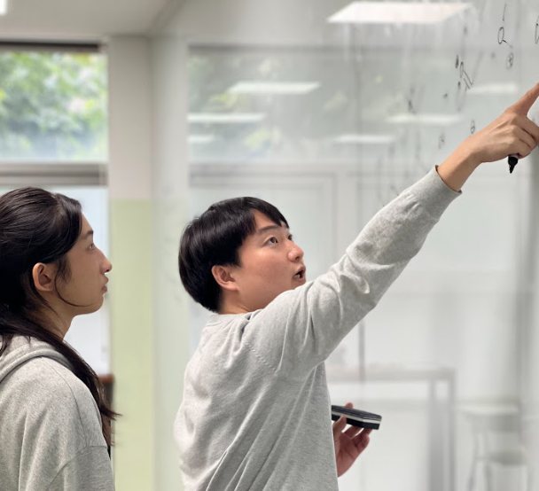 A young man in a gray sweater points to handwritten notes high on a whiteboard while a young woman beside him follows along
