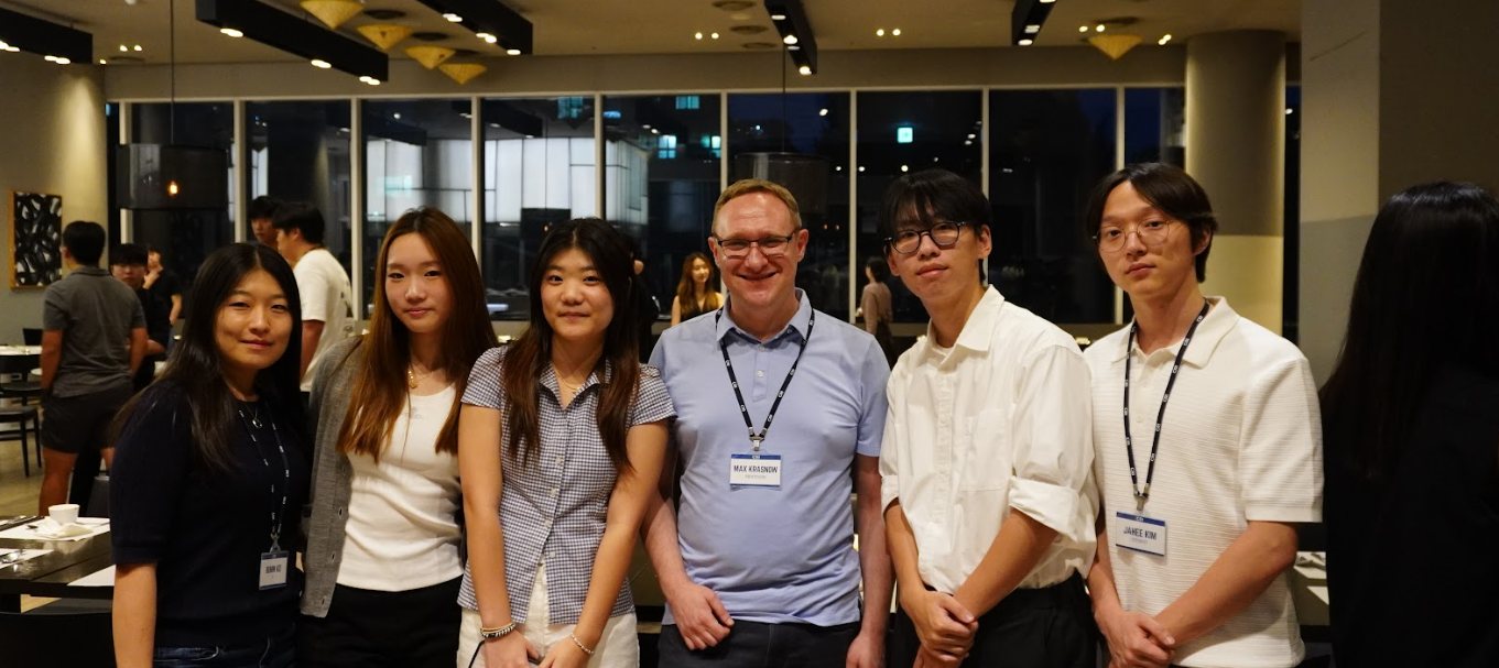 Professor Max Krasnow, wearing a name badge, stands with five others at an evening event in a room with tall windows