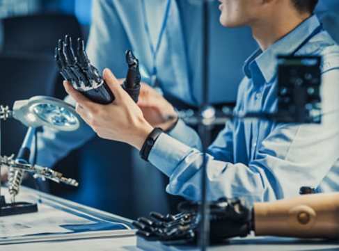 Two people at a lab workbench examine a black robotic prosthetic hand, with a prosthetic arm and tools nearby