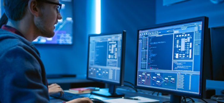 A programmer in glasses works at two monitors showing code and system diagrams in a blue-lit room
