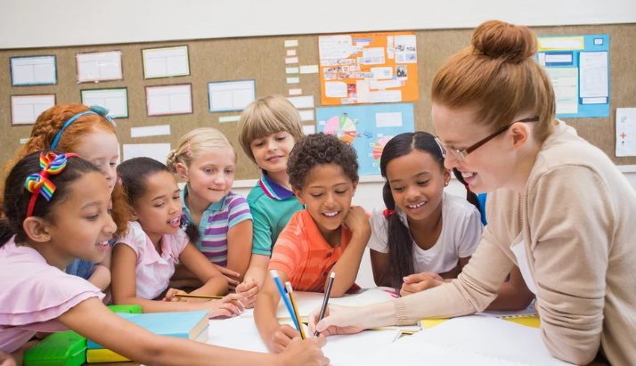 A teacher leans over a table of smiling young children writing with pencils in a primary school classroom