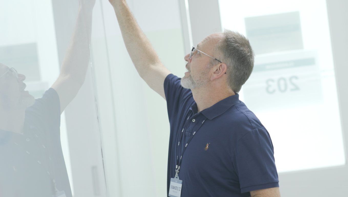 Brian Stoltz reaches up to write on a whiteboard in a bright classroom
