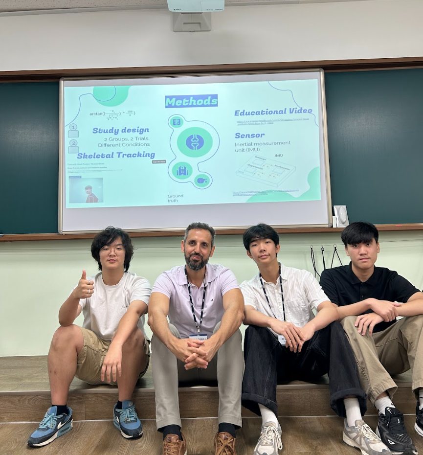Professor Alshurafa sits with three students in front of a projected slide listing study design, skeletal tracking, and an inertial measurement unit sensor
