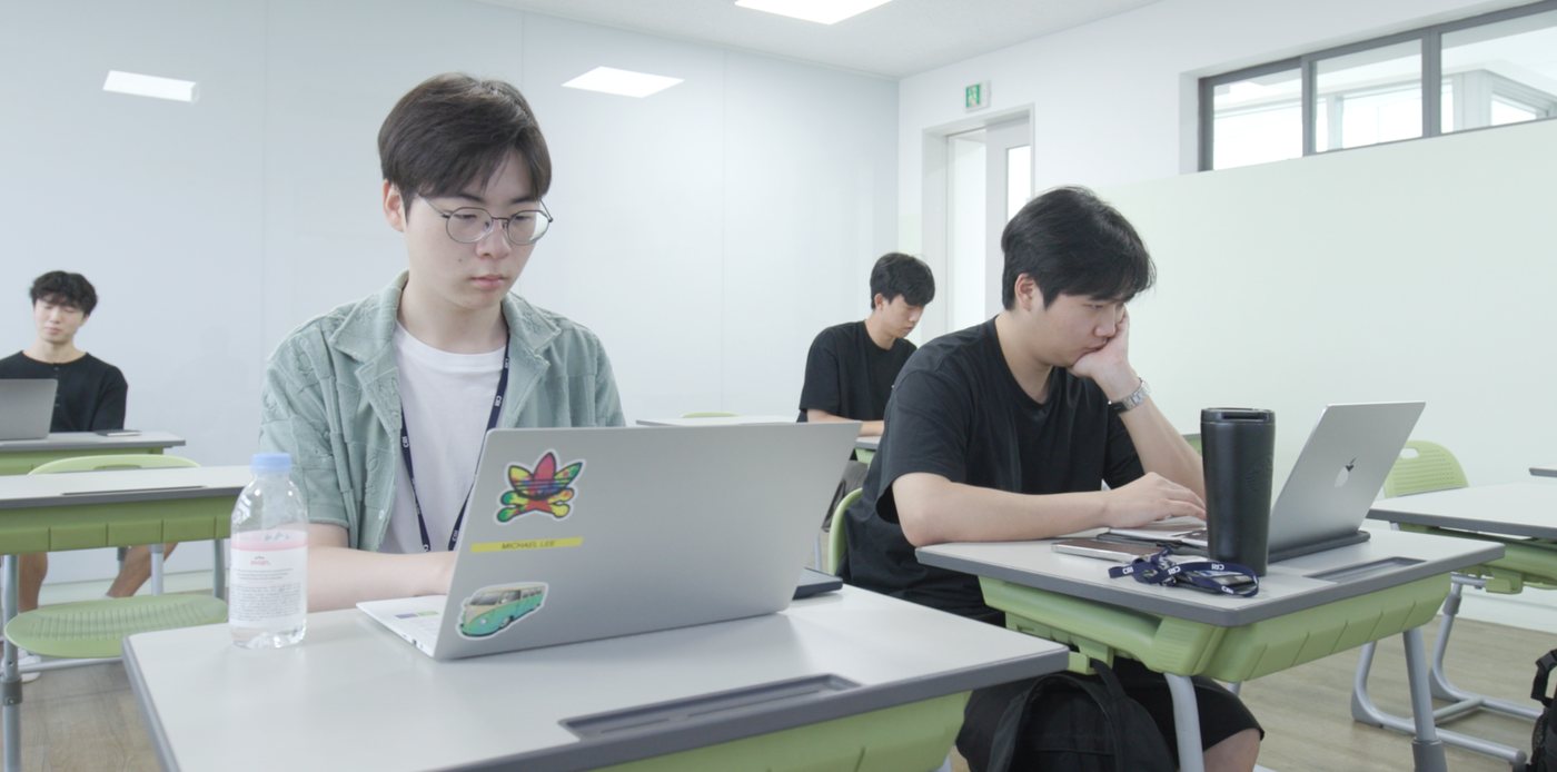 Students work on laptops at desks in a bright classroom