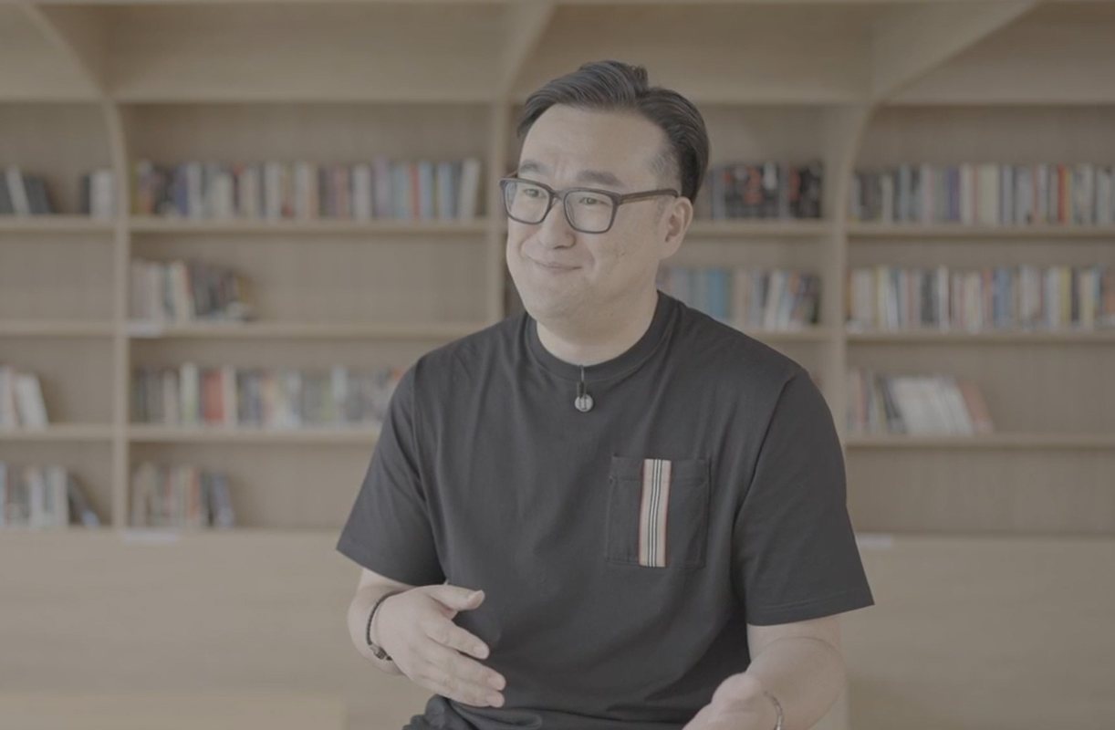 Ryan Fang, wearing glasses and a black T-shirt, gestures as he speaks in front of wooden bookshelves