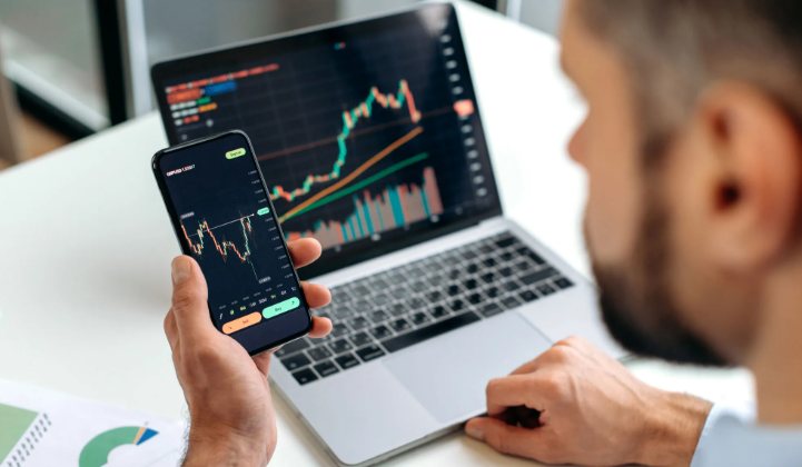 A hand holds a smartphone showing a candlestick price chart in front of a laptop displaying stock charts