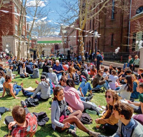 Students sit on a sunny campus lawn beneath a "Lawn Party" banner, with brick buildings behind them