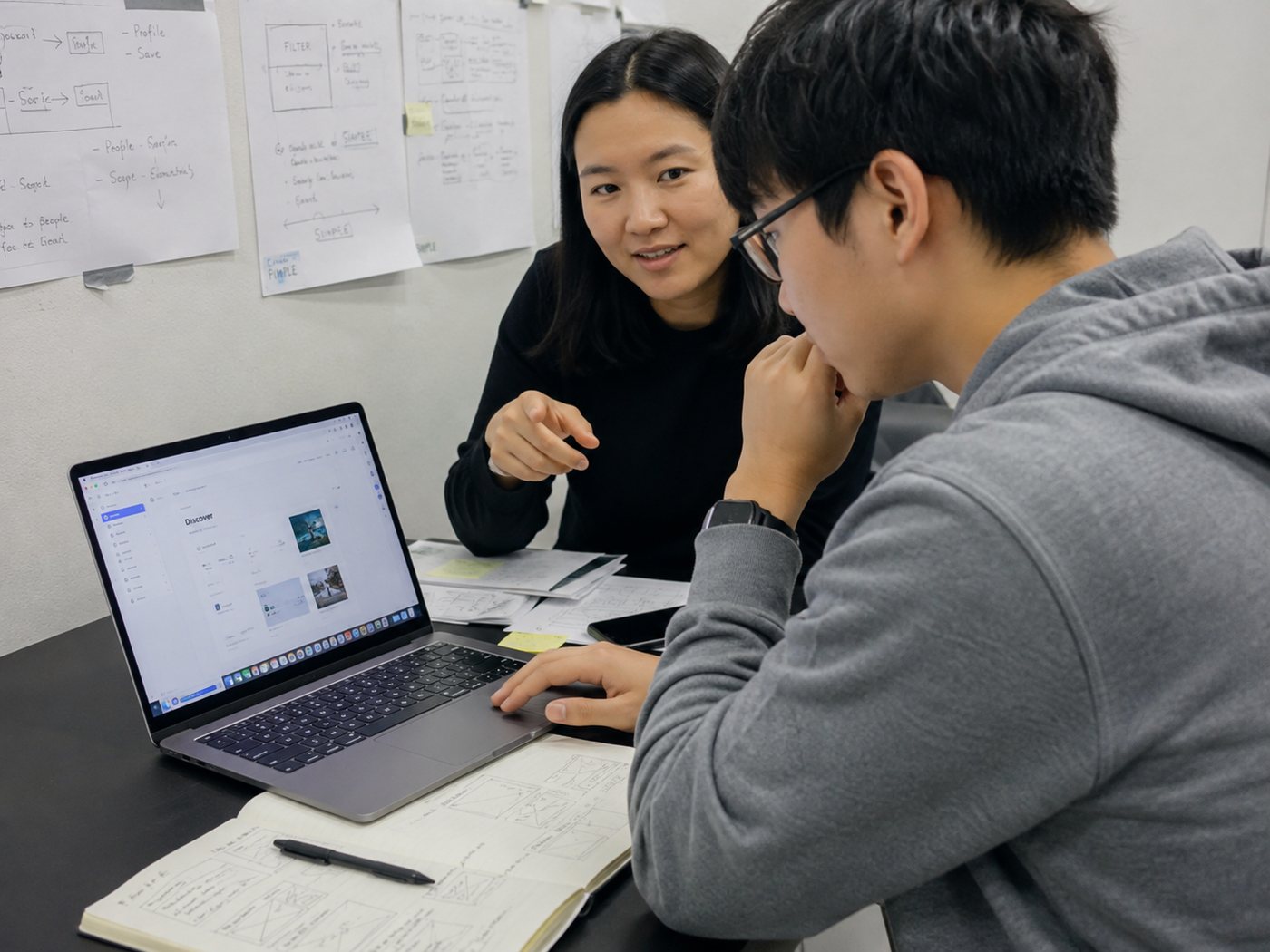 Illustration of a mentor gesturing toward a laptop as a student in glasses studies the screen, with sketches pinned to the wall behind them
